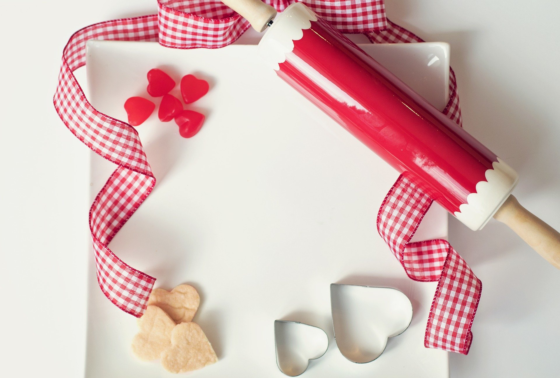 A red and white gingham ribbon on a plate, with a red and white rolling pin, heart shaped sweets, 2 heart cookie cutters and 3 hearts shaped cookies.