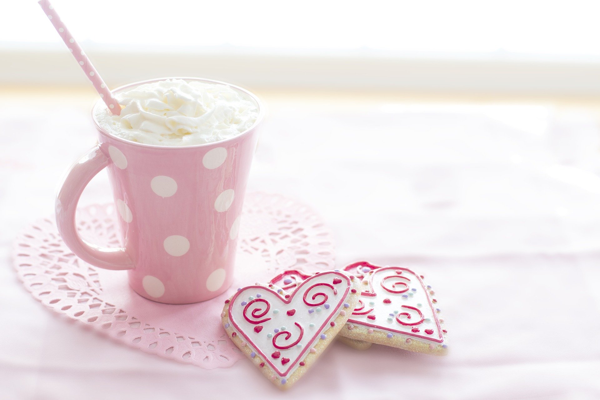 A pink mug with white polkadots with with cream and a pink and white spotted straw. It is sitting on a pink heart shaped mat, with 2 heart shaped iced cookies next to it.