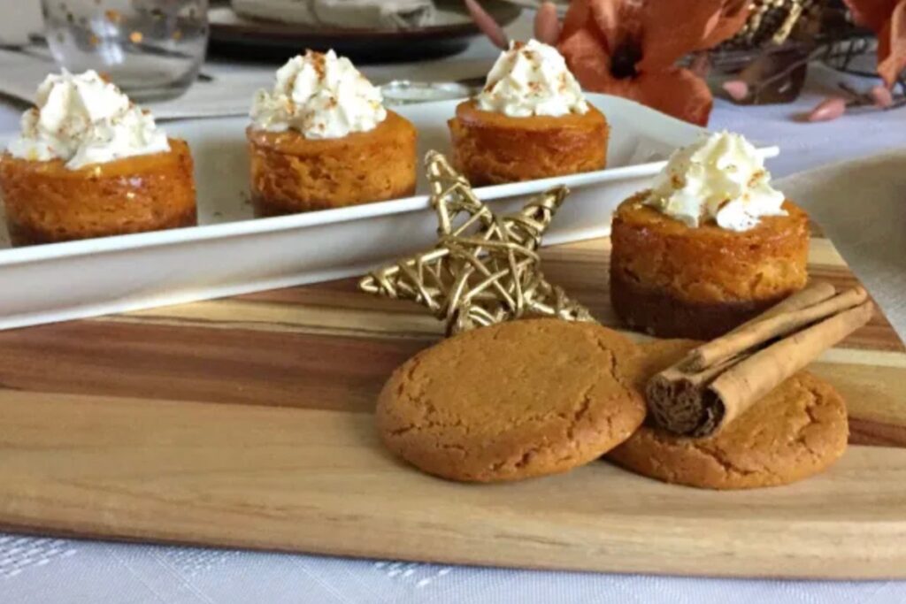 Mini pumpkin cheesecakes on a white platter. In front is a wooden board with a single cheesecake on it, a cinnamon stick, a golden star and 2 cookies
