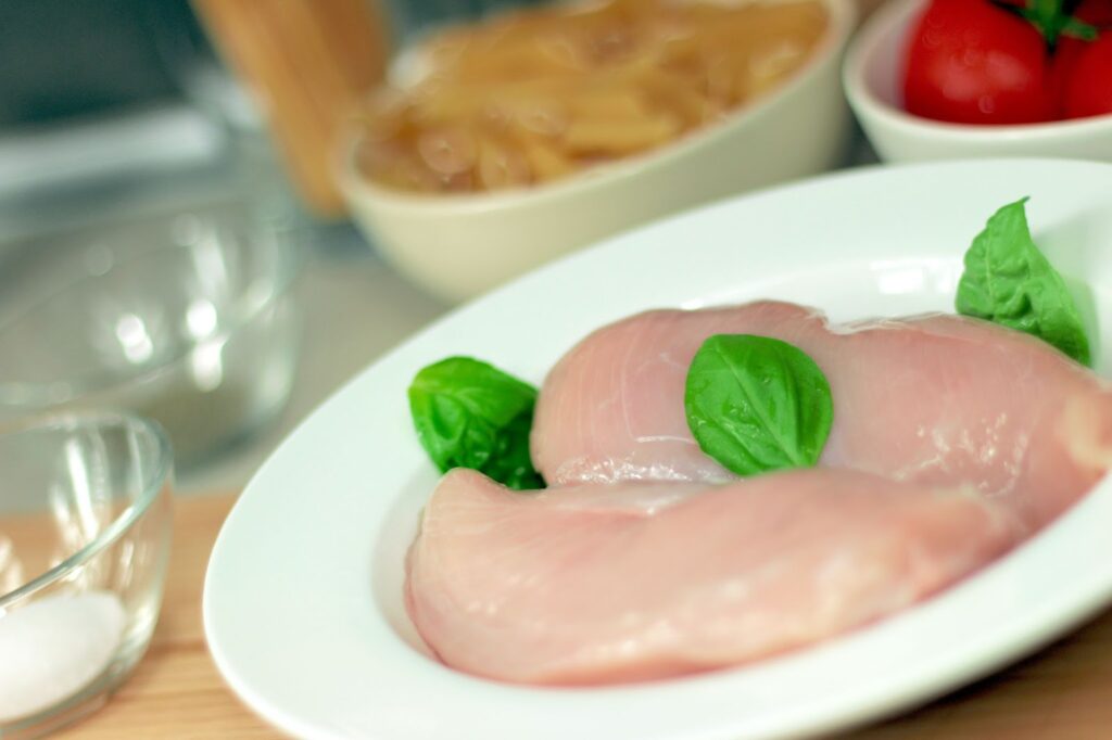 A dish with raw chicken and basil on a plate. Around it and slightly out of focus is a bowl of raw pasta and a bowl of tomatoes.