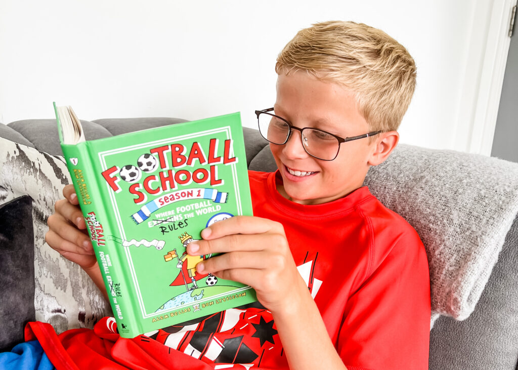 A boy reading a book called "Football School". He has blonde hair and glasses and is wearing a red t-shirt