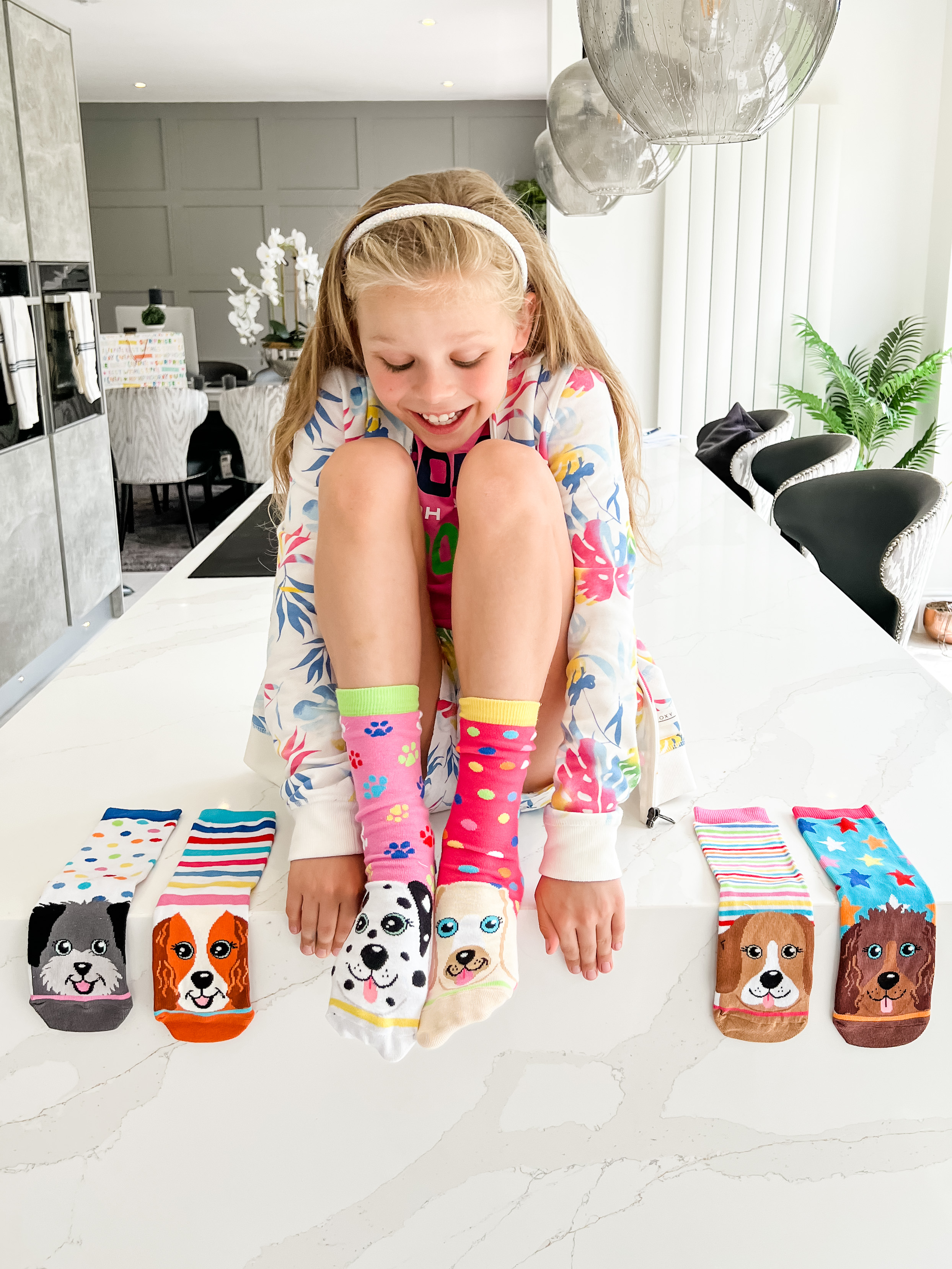 A girl sitting on the edge of a counter. She is wearing odd shocks with dogs on the toes, with 4 other odd socks with dogs on them lined up on either side of her.