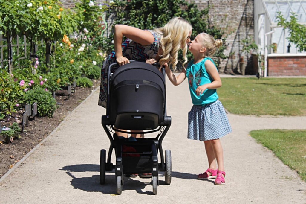 A woman pushing a black pram. She is bending down to kiss a young girl standing next to her