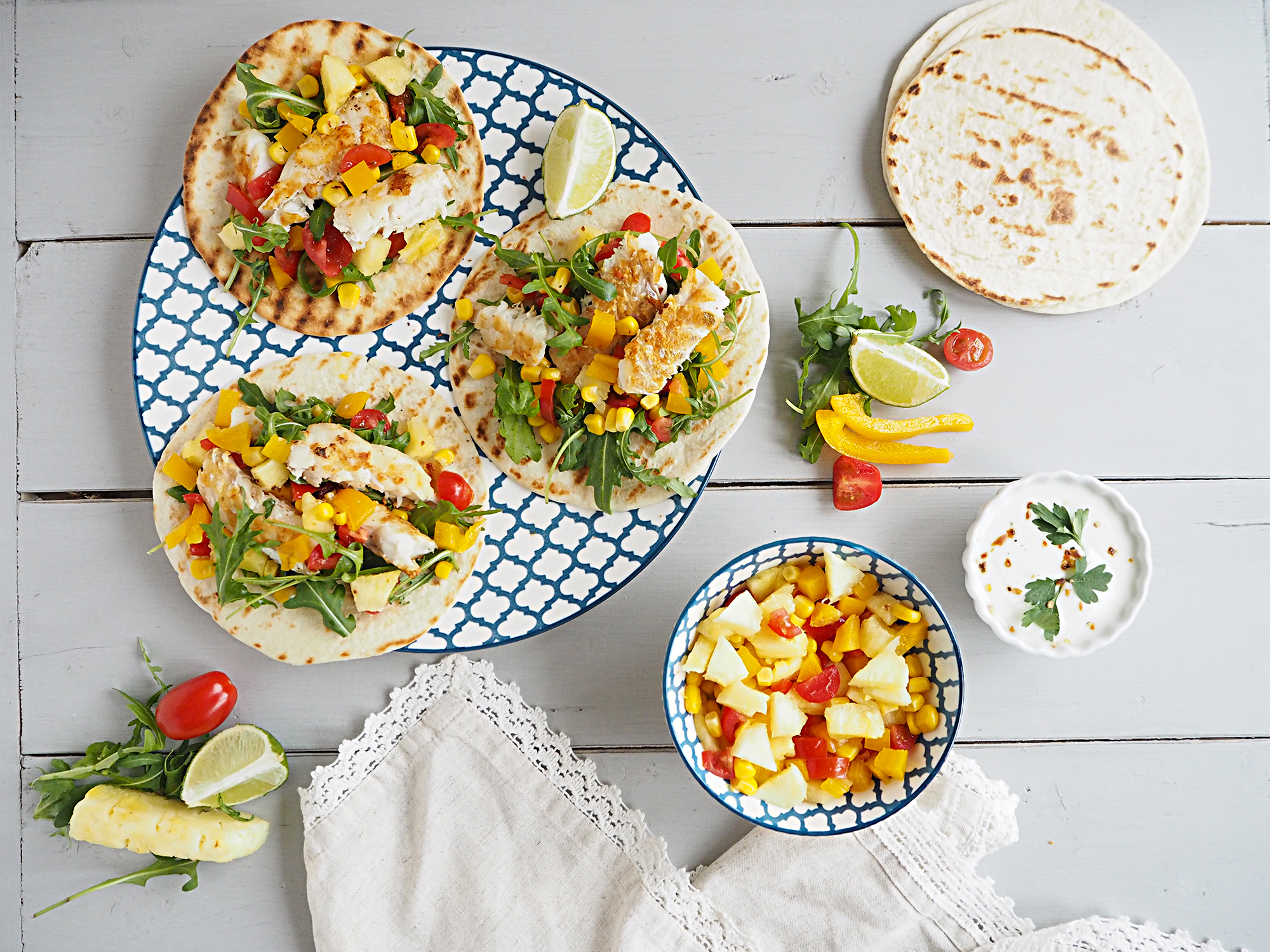 A plate of fish tacos with pineapple salsa, next to a bowl of salsa, more tortillas and some natural yoghurt.