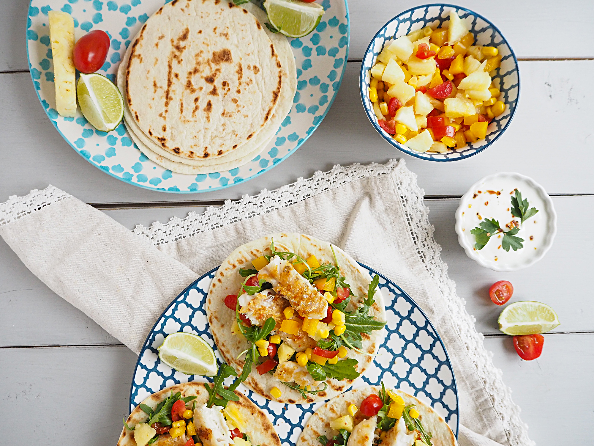 A blue and white patterned plate sits on a white napkin. On the plate are 3 open fish tacos with pineapple salsa. Above this is a bowl of pineapple salsa and a plate containing more tortillas.