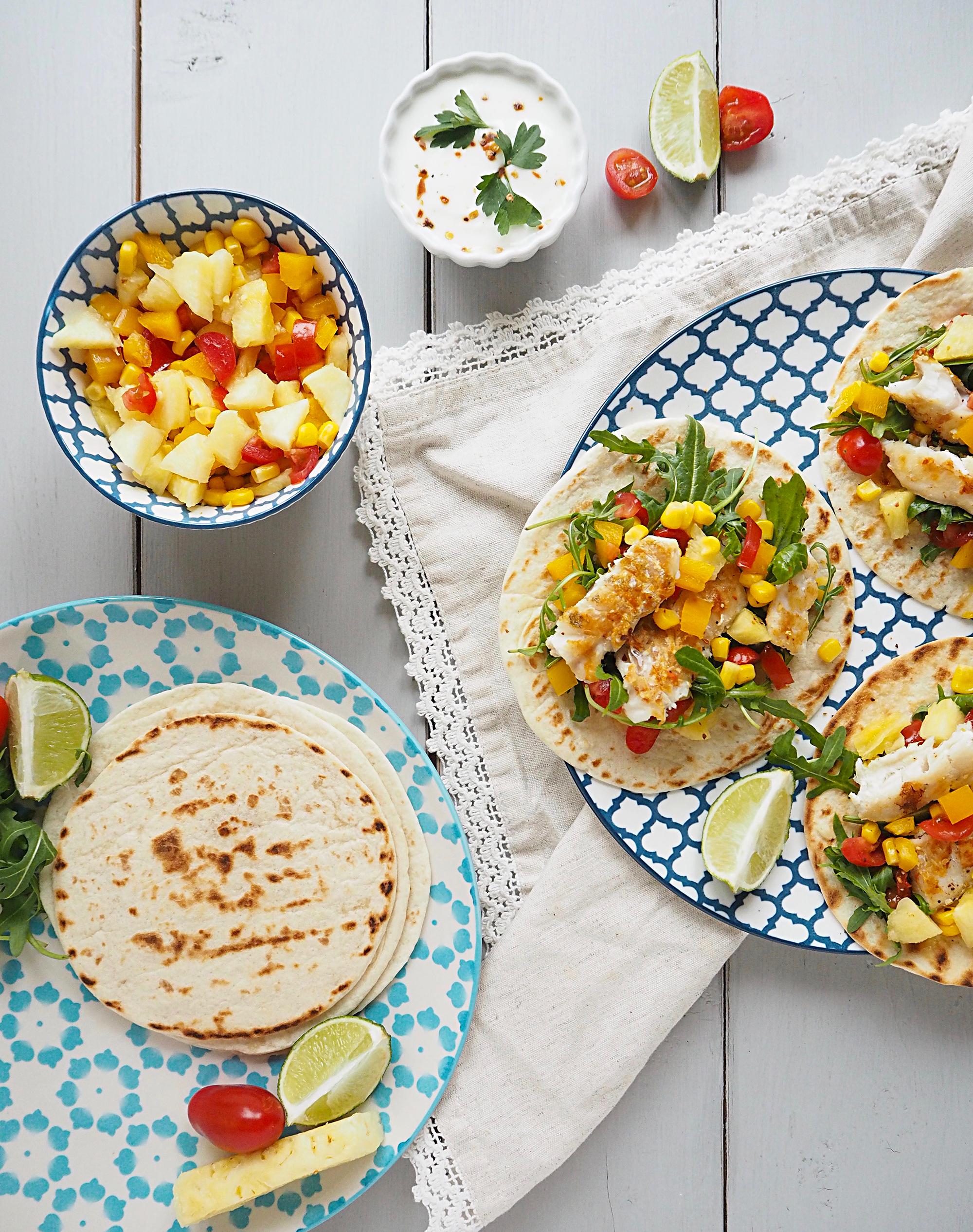 A blue and white patterned plate sits on a white napkin. On the plate are 3 open fish tacos with pineapple salsa. To the left is a bowl of pineapple salsa and a plate containing more tortillas.