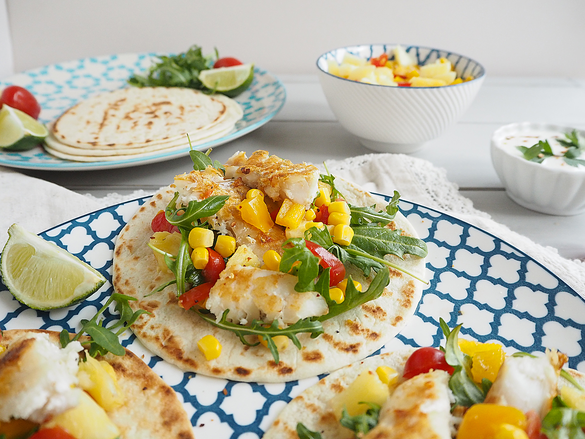 A plate with Fish Tacos with Pineapple Salsa on it, next to more plates containing tortillas, more salsa and yoghurt.