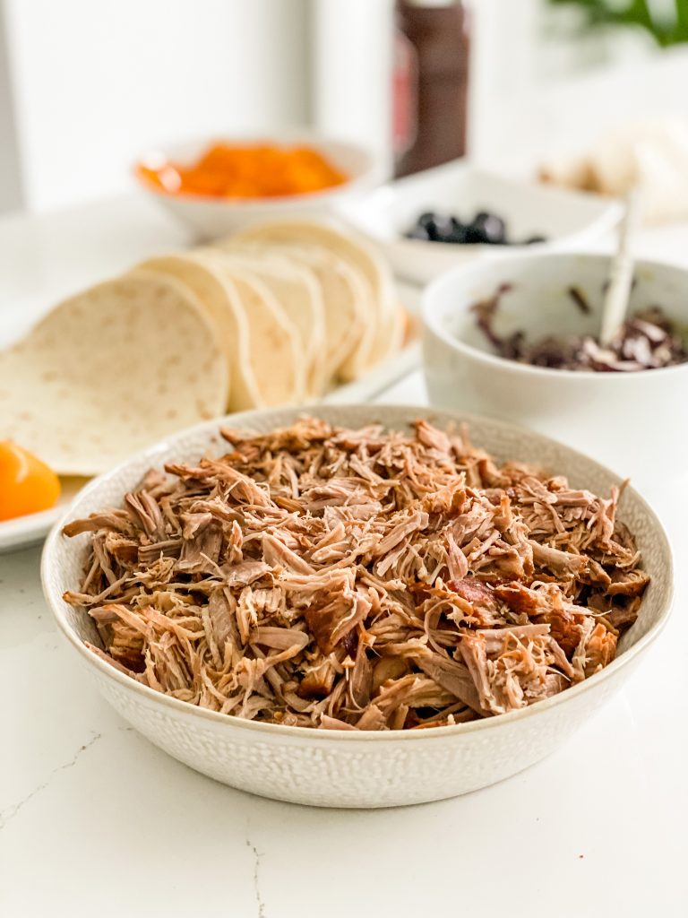Shredded pulled pork in a while dish. Behind is a plate of tortillas and some slaw