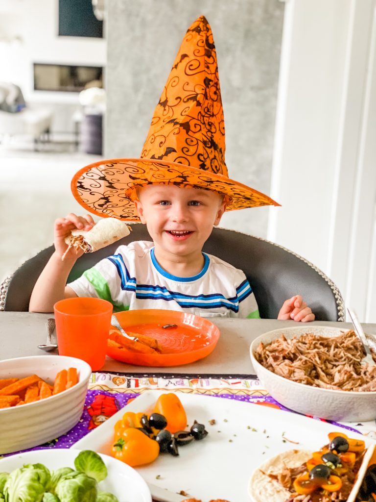 A little boy in an orange and black Halloween hat smiling and holding a taco