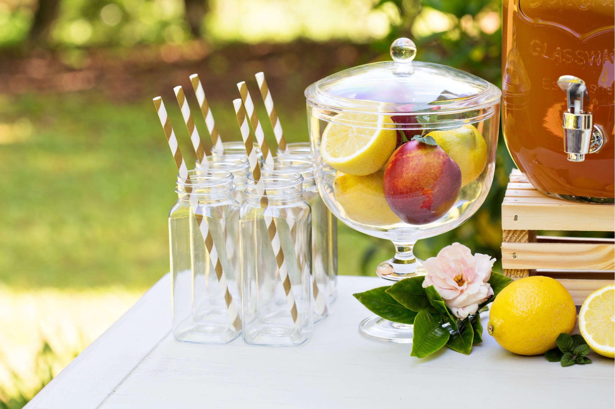 A table set up on the grass. It has glasses with striped straws on it, a bowl of fruit and a drinks dispenser fill with some sort of punch
