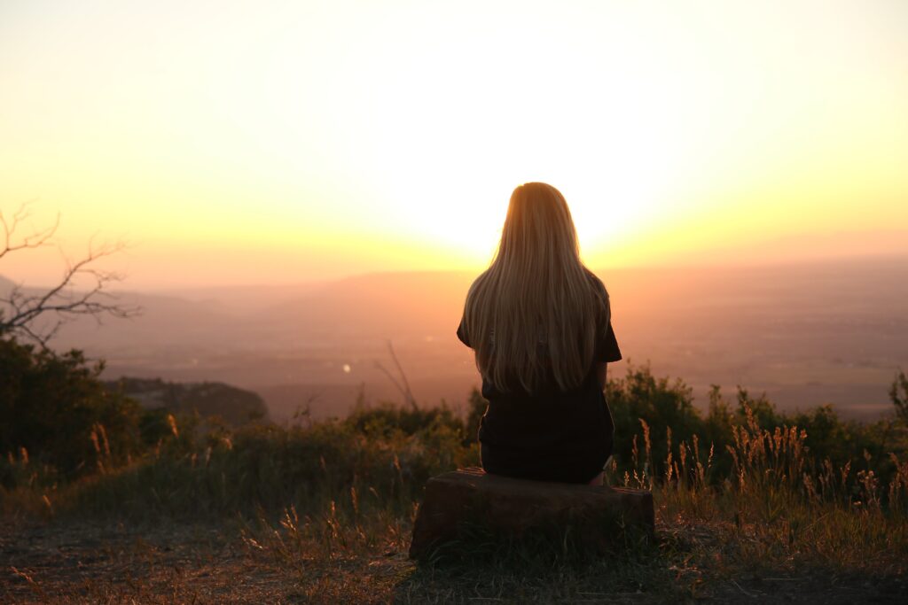 A woman sitting on top of a hill watching the sunset, feeling serene and appreciative of the natural beauty around her.