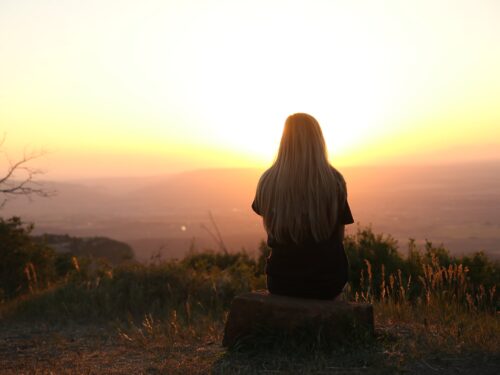 A woman sitting on top of a hill watching the sunset, feeling serene and appreciative of the natural beauty around her.