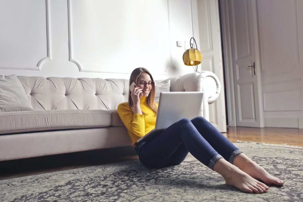 A young woman sitting on the cozy flooring with a laptop in front of a couch in her family home.
