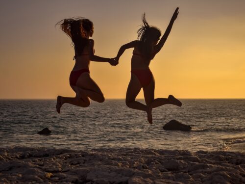 Two women jumping in the air on the beach at sunset, exemplifying a summer body.