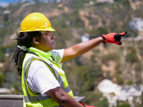 A woman wearing a hard hat and safety vest pointing to a hillside.