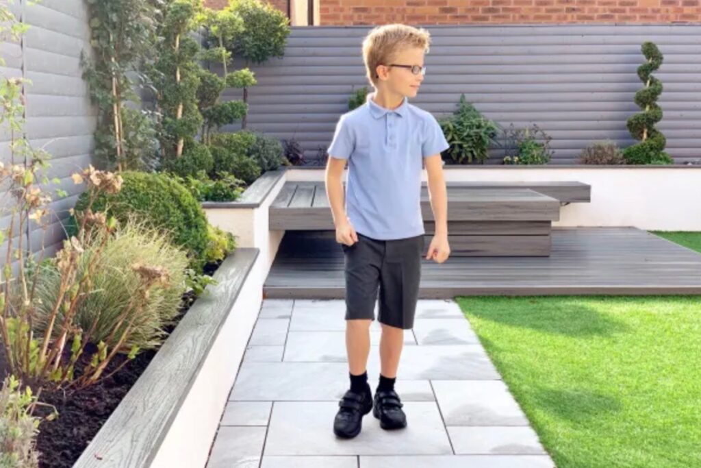 A boys standing on a garden patio in grey school shorts and a blue polo shirt with black school shoes