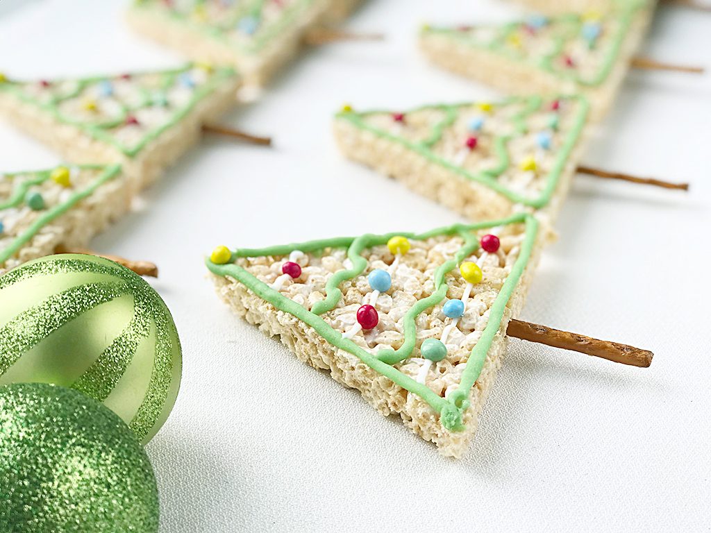 Rice Krispy Treats Christmas Trees with pretzel trunks lines up, with 2 glittery green baubles next to them