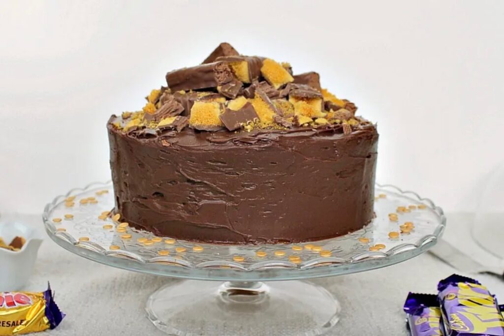A cake covered in chocolate icing and topped with crushed Crunchie bars and twirls. It sits on a glass cake stand.