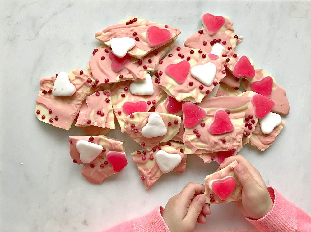 A childs hand holding a piece of chocolate bark with hearts and sprinkles on it for Valentine's Day. There is a big pile of bark next to her.