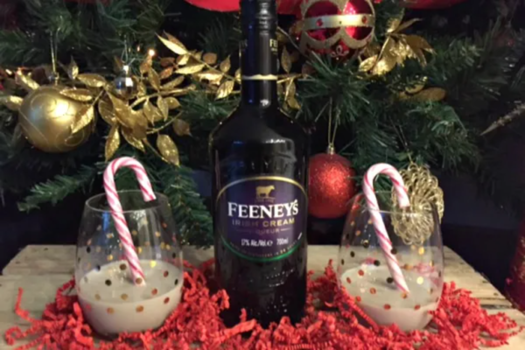 A bottle of irish cream in front of a Christmas tree. 2 glasses are on either side of it with Irish cream and a candy cane in them