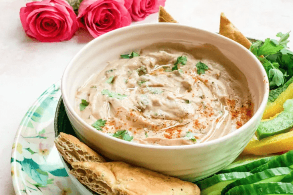 A bowl of pink coloured baba ganoush on a plate with bread and vegetables