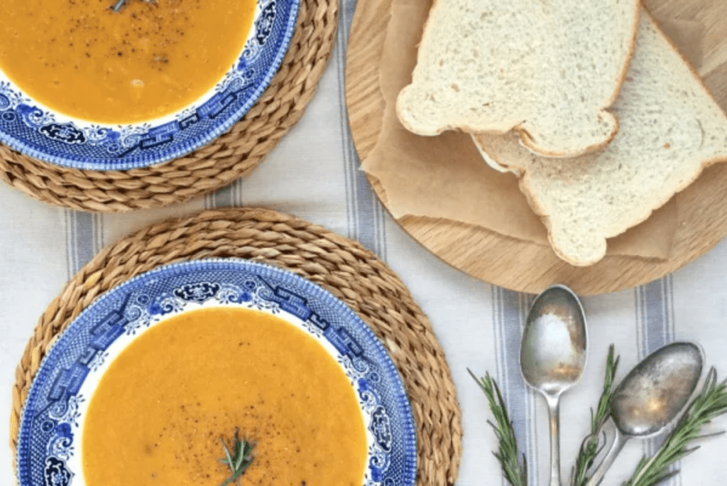 2 bowls of butternut squash soup in blue bowls on woven place mats. 2 slices of white bread are on a wooden board next to them