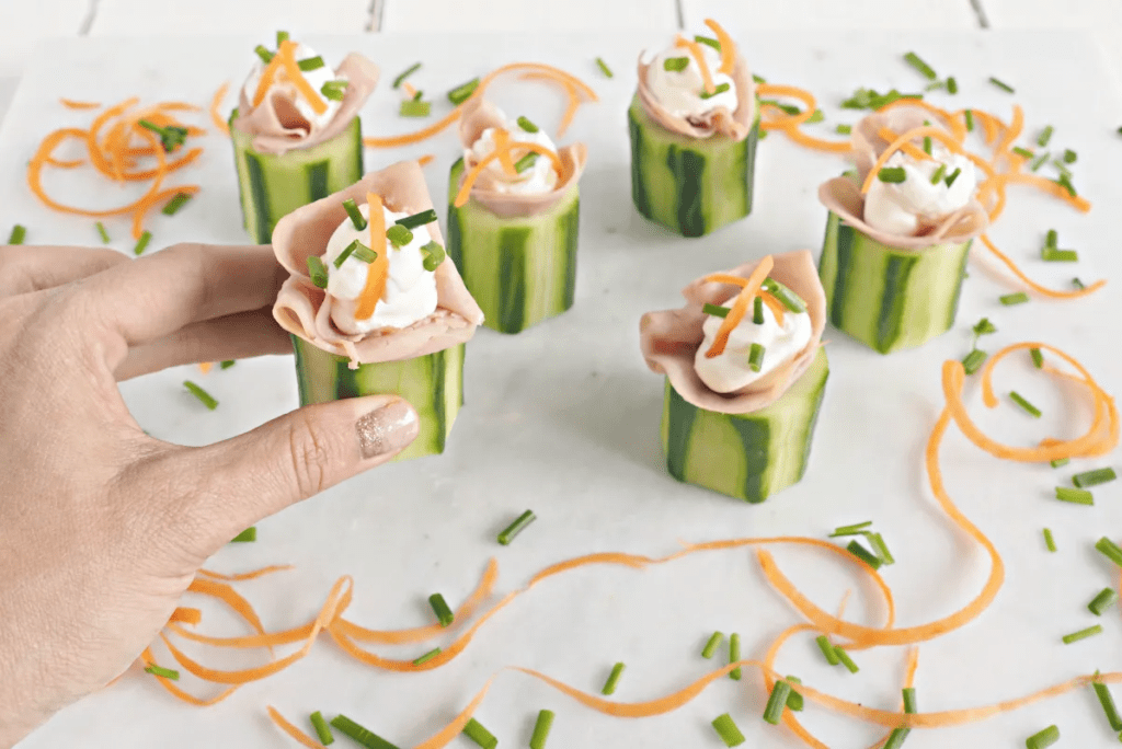 Chunks of cucumber topped with ham and cream cheese. A hand is picking one of them up.