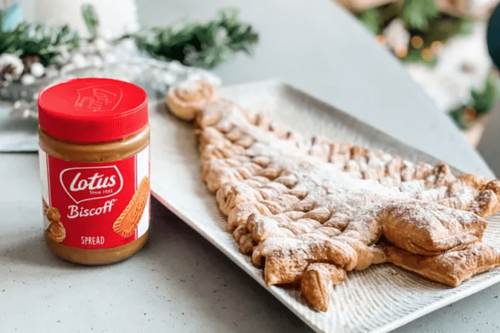 A jar of Lotus Biscoff spread next to a plate. On the plate is a Christmas Tree shape made out of puff pastry, and sprinkled with white icing sugar