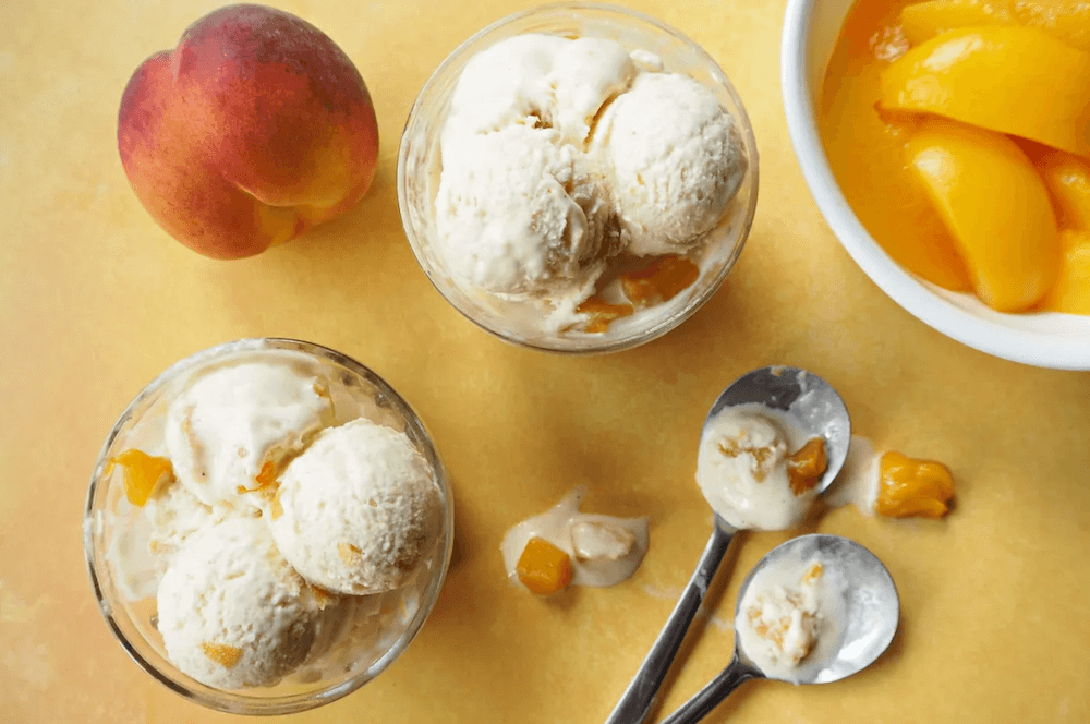 Two bowls filled with peach ice cream. They are set on a yellow background and accompanied by a bowl of sliced peaches, a whole peach and 2 spoons with slightly melted ice cream on them 