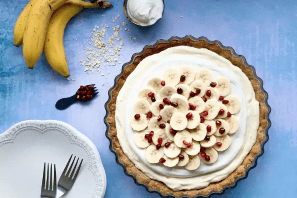 A banana cream pie on a blue counter. Next to it is a bunch of bananas and a plate with 2 forks on it