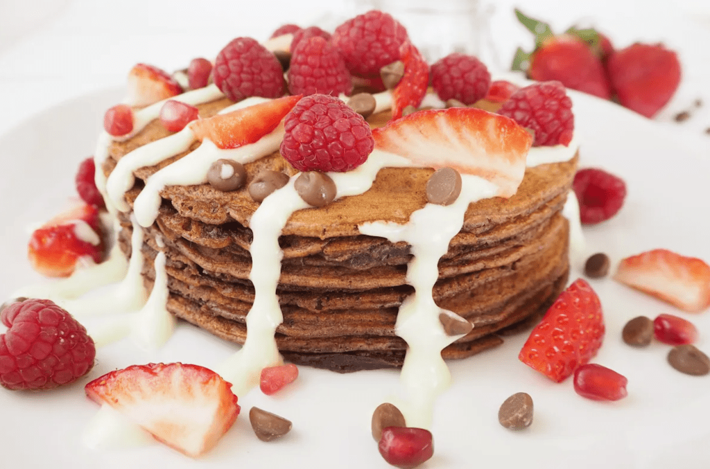A stack of chocolate pancakes drizzled with cream and topped with chocolate chips and red berries