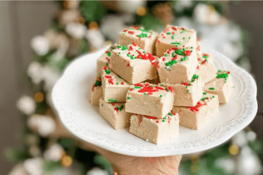 Peppermint White Chocolate Fudge topped with red and green sprinkles. it is on a white plate and being held infront of an out of focus Christmas tree
